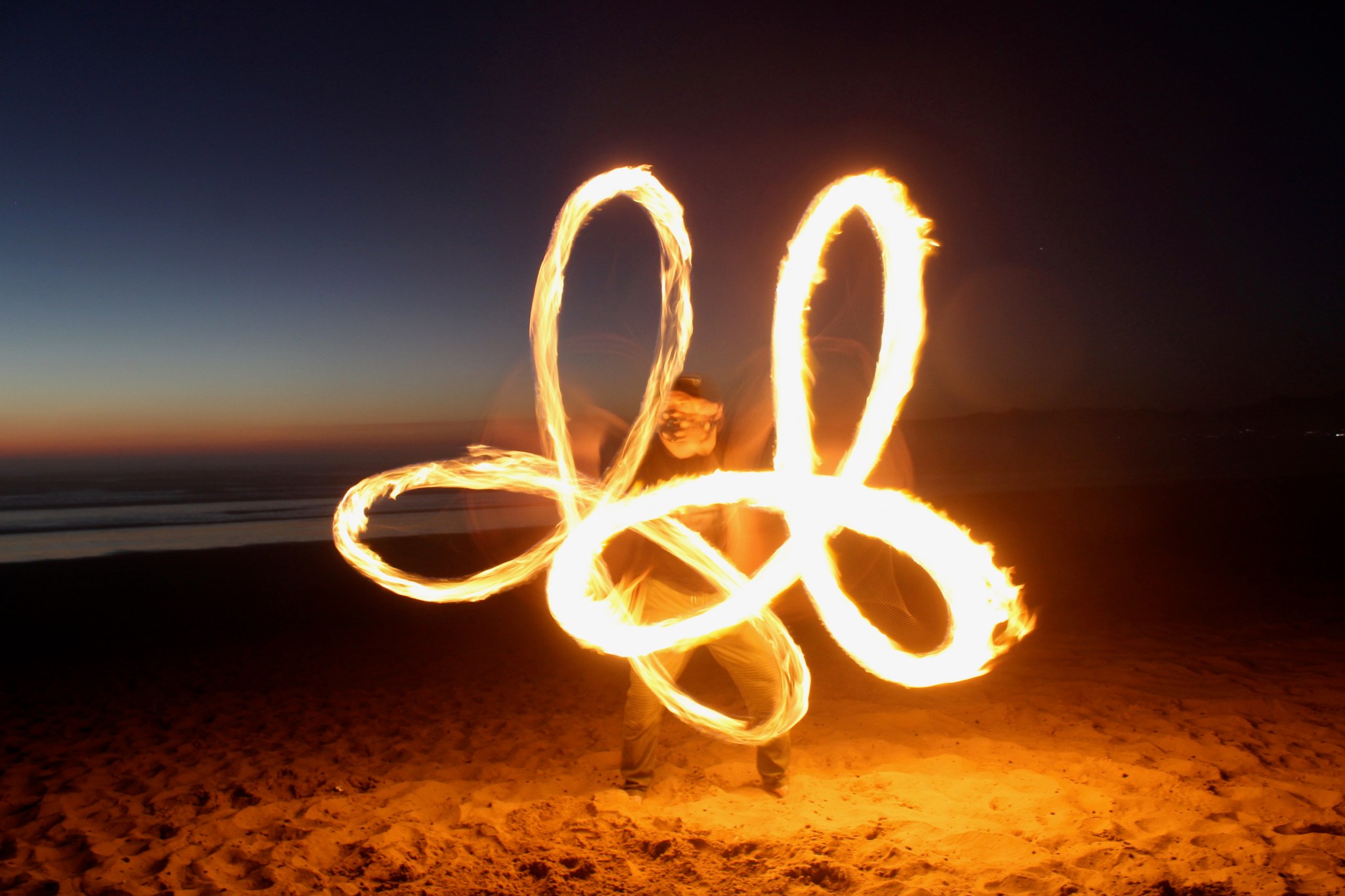 A long-exposure photo of a man fire spinning on a beach, creating a geometric shape with the flames.