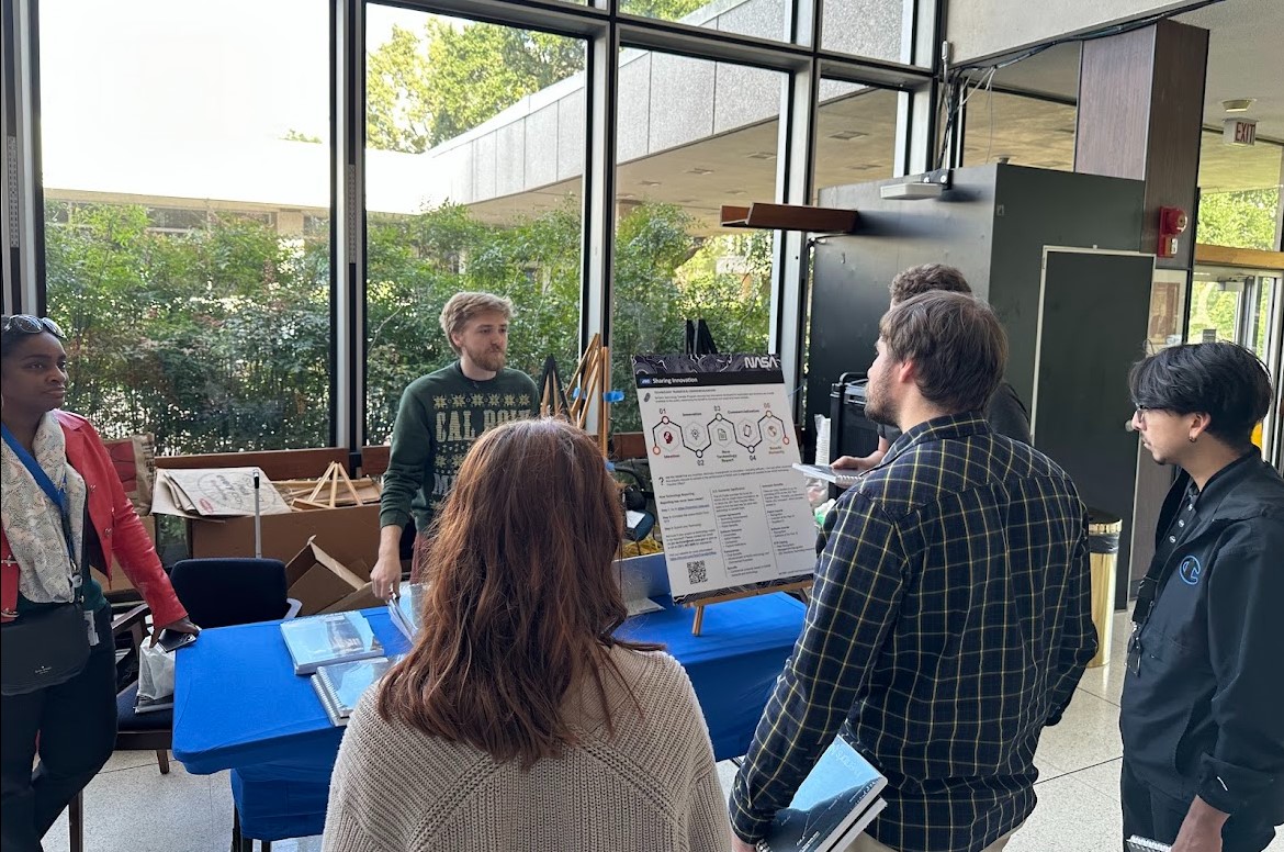 A man wearing a holiday sweater stands behind a table, presenting information about technology transfer to a group of his peers.