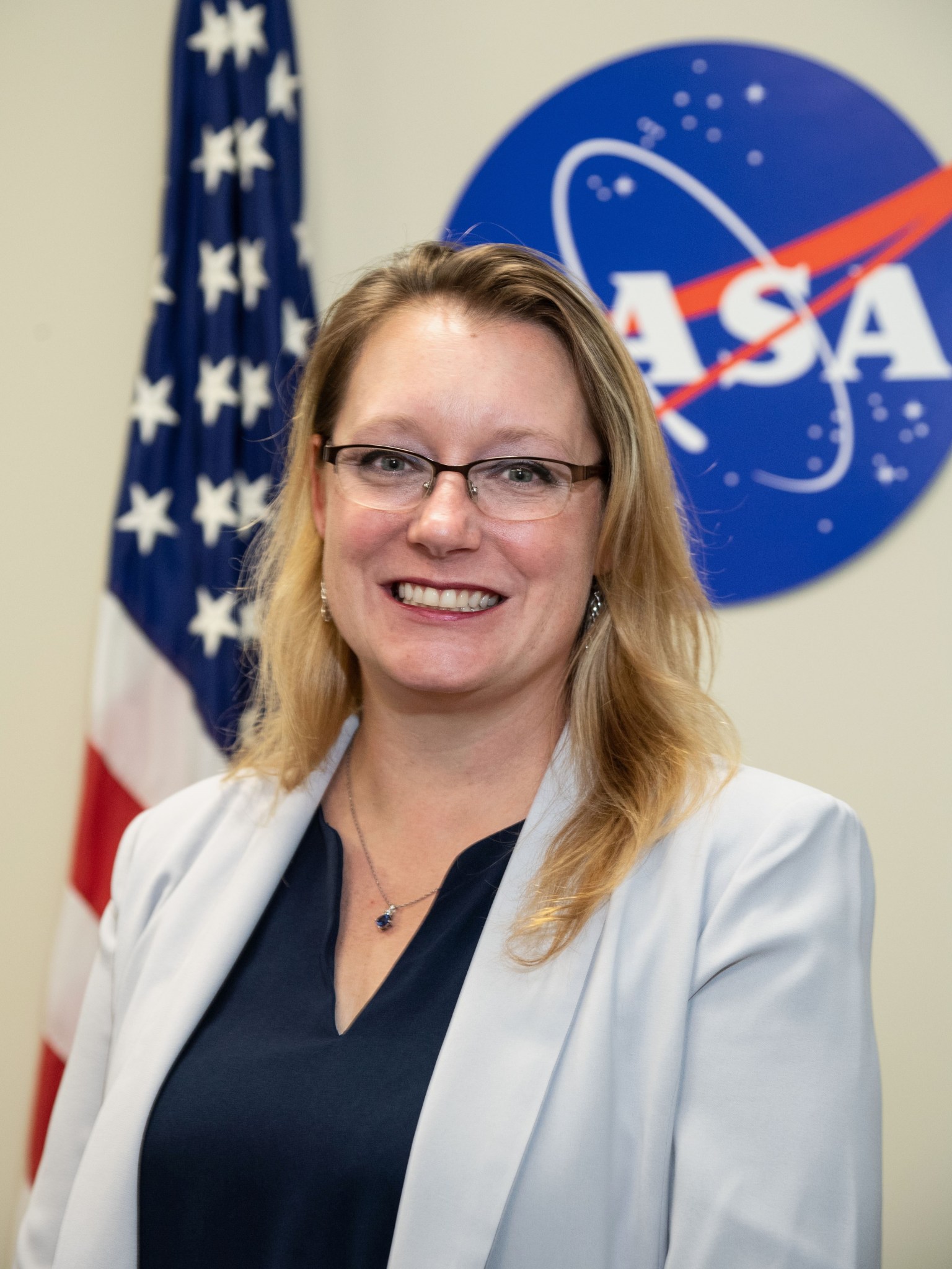 A woman in a cream blouse and black T-shirt poses in front of a NASA meatball logo (right) and the U.S. flag.