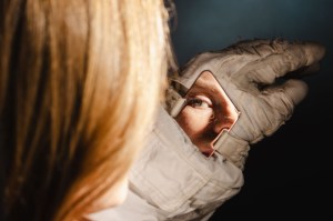 A white woman's eye and nose are reflected in a diamond-shaped mirror attached to the wrist of her spacesuit. The mirror and white spacesuit glove are in focus, while the astronaut's blonde hair is in the foreground, out of focus.