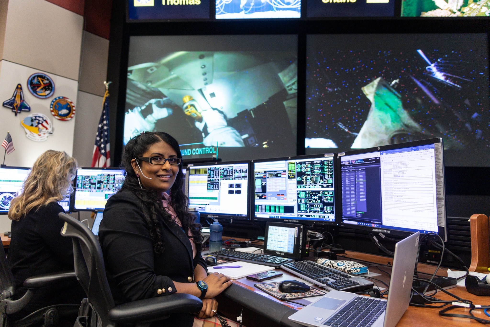A woman sits at a desk and poses in front of three monitors. Multiple monitors display live data and video of astronauts on the International Space Station. An American flag and NASA mission patches are visible on the wall behind her.