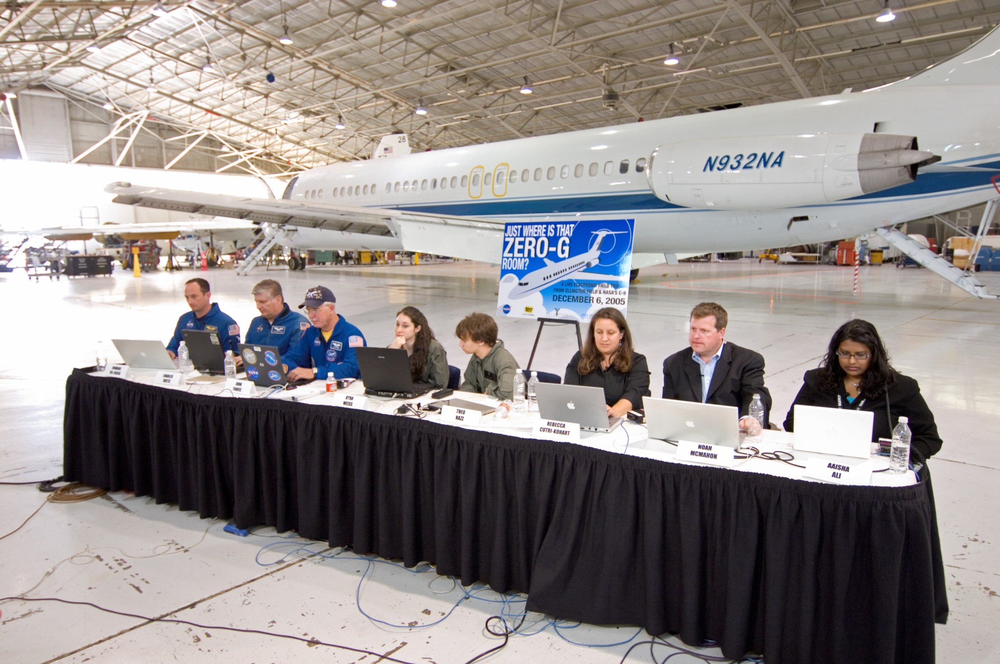 Eight people sit at a long table in front of their computers in a large facility. An airplane is parked behind them.