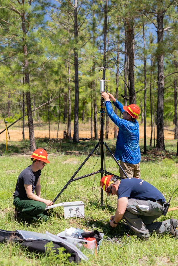 Three men in blue shirts with red hats mount equipment on tripod in grassy field.