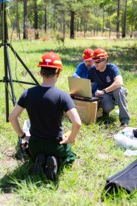 Three men in blue shirts with red hats mount equipment on tripod in grassy field.