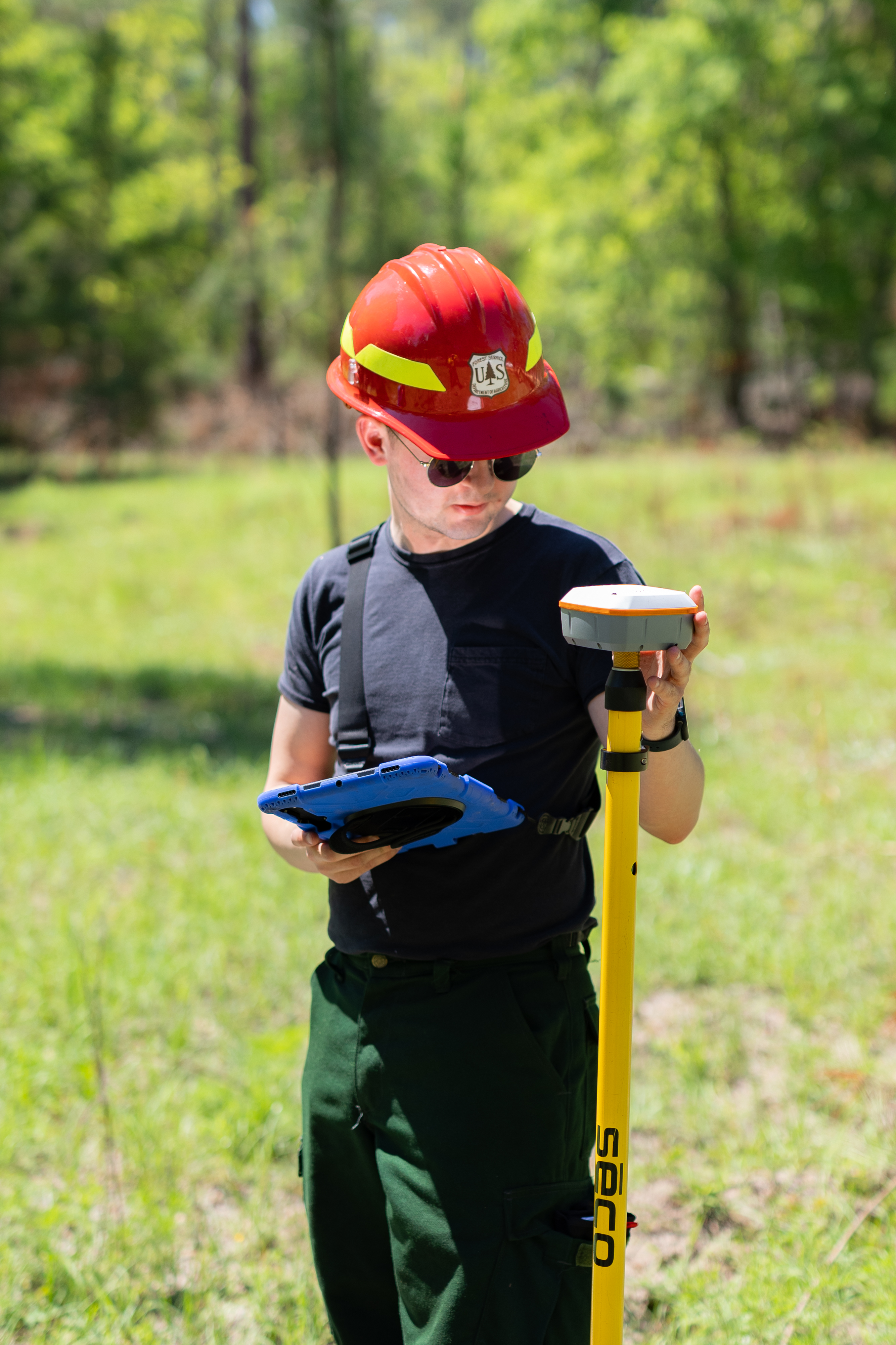 A young man in a red hard hat holds a tablet and checks a gps unit in forest clearing.