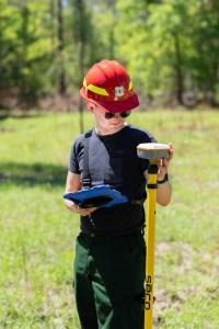 A young man in a red hard hat holds a tablet and checks a gps unit in forest clearing.