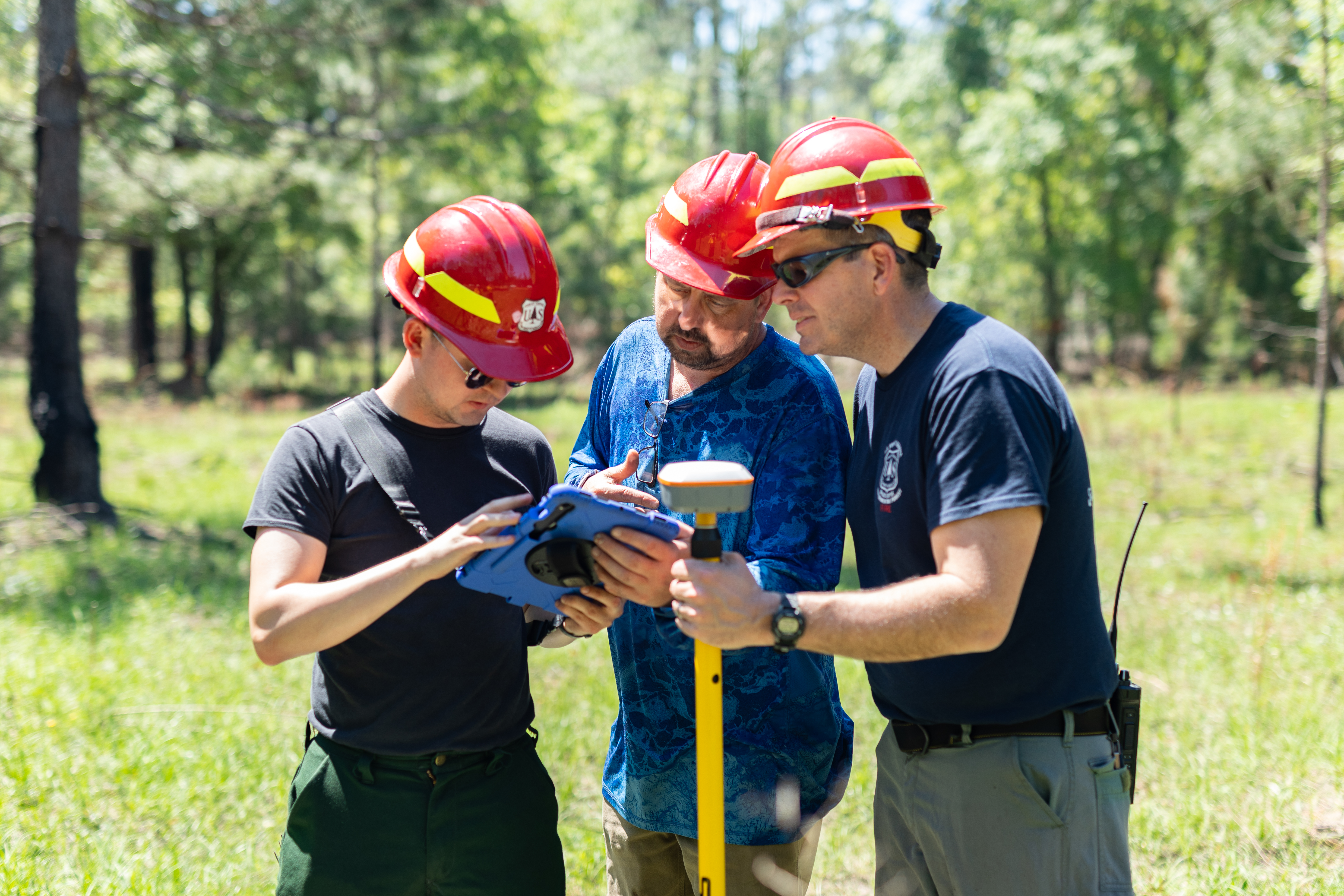 Three men in red hardhats look at an ipad in grassy field, while the one on the far right hold a gps unit on a pole.