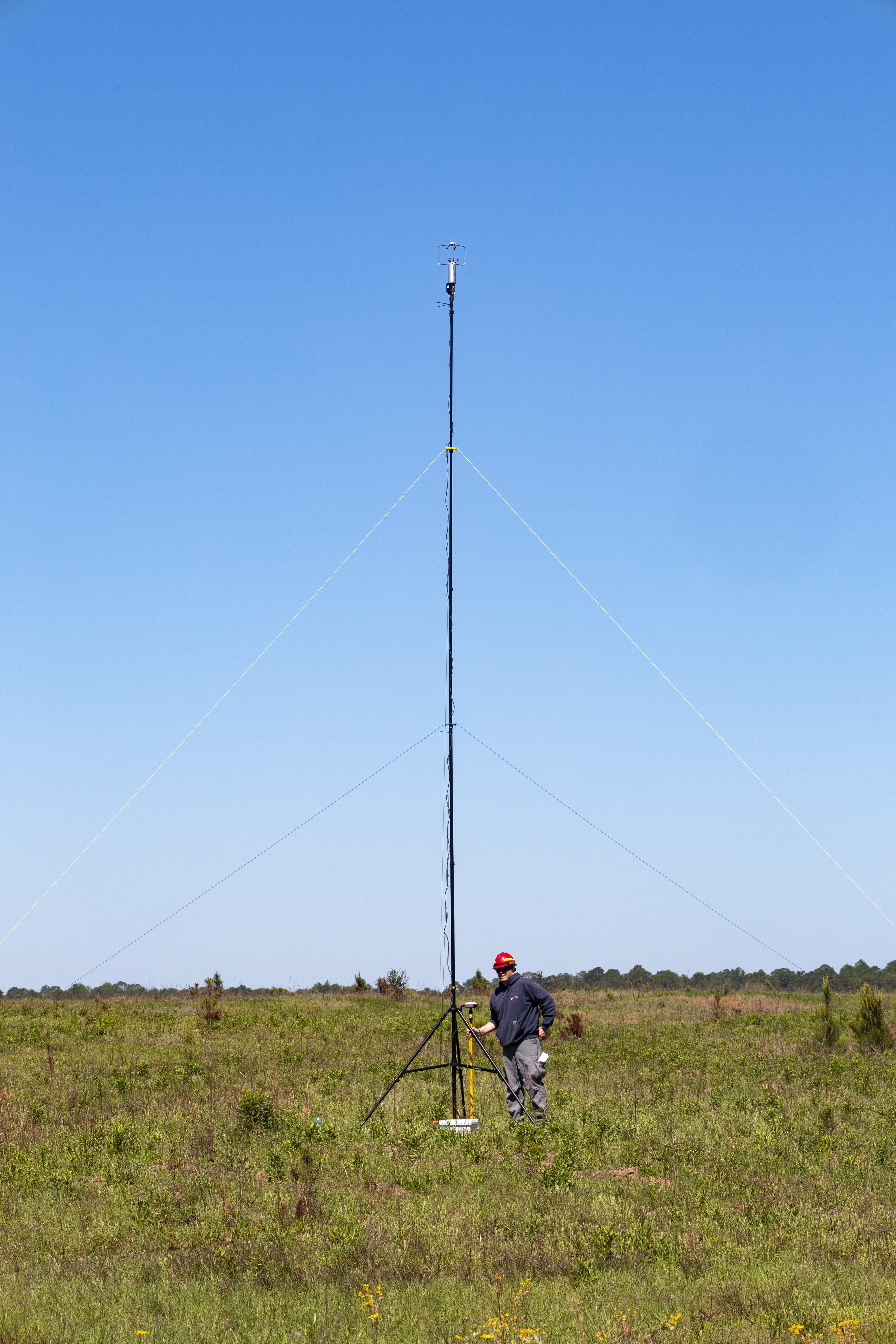 A man in a navy long-sleeved and red hard hat checks equipment mounted on a tripod in a grassy field.