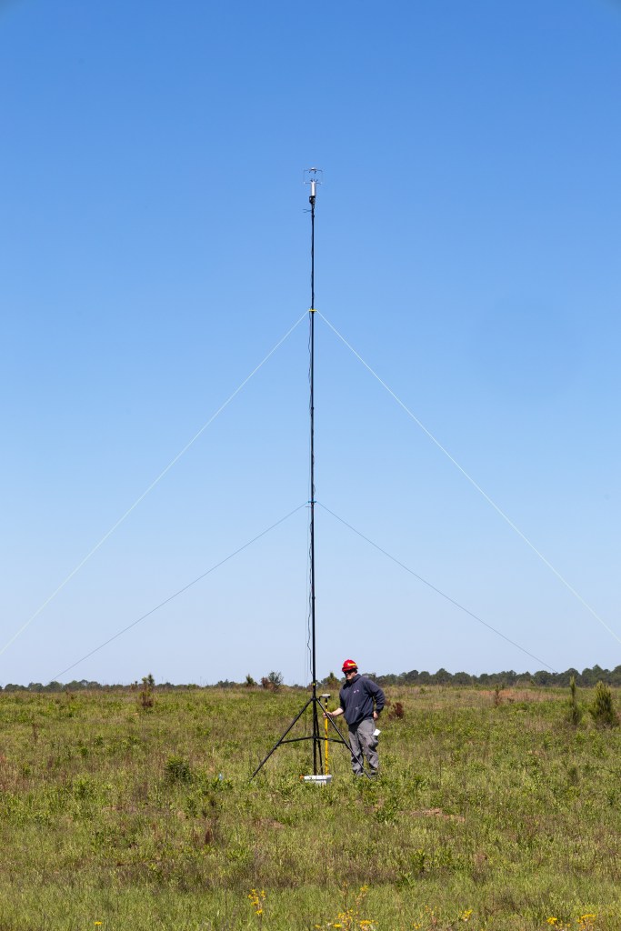 A man in a navy long-sleeved and red hard hat checks equipment mounted on a tripod in a grassy field.