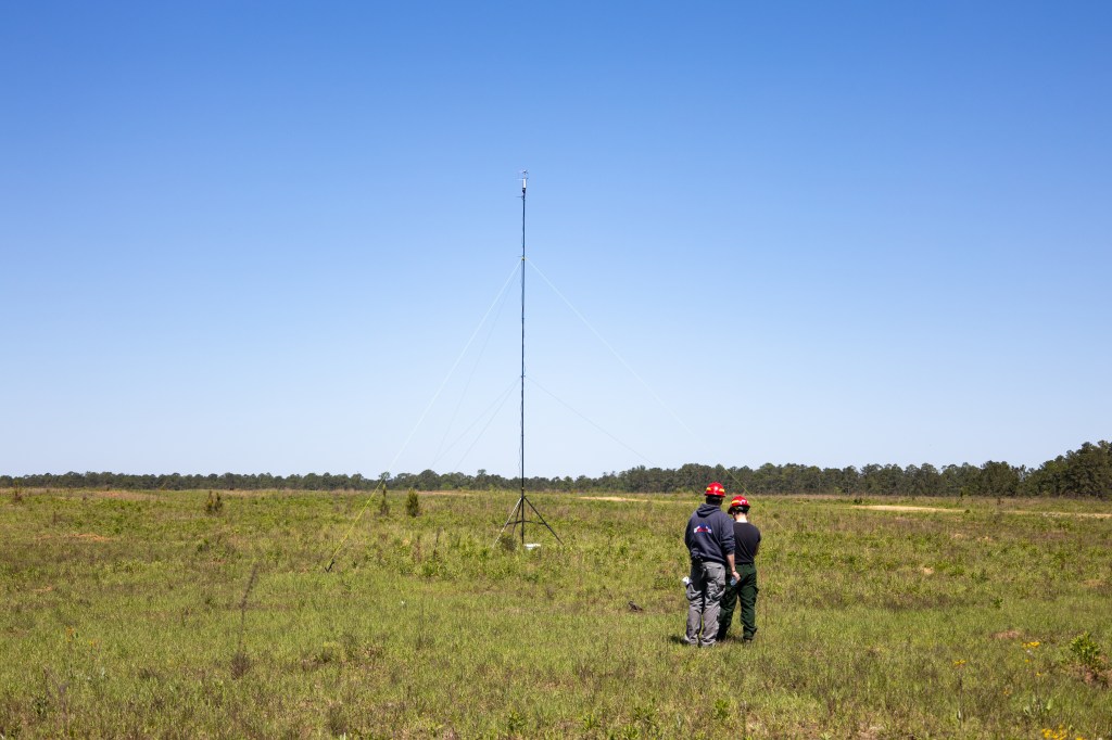 Two men in navy blue with red hardhats stand with their backs to the camera in a grassy field. In front of them is a piece of equipment mounted on a large tripod.