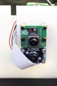 Two tiny camera lenses are mounted on a piece of white plastic, with a small red wire coming out the side.