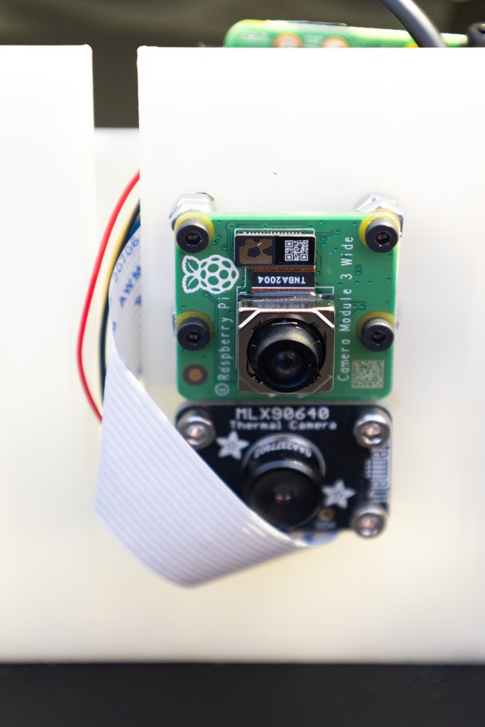 Two tiny camera lenses are mounted on a piece of white plastic, with a small red wire coming out the side.