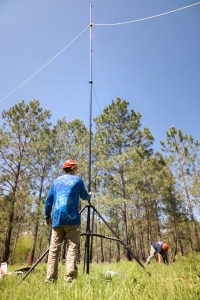 Two men in blue shirts with red hardhats mount equipment secure long, thin lines to support equipment on a tall tripod in a grassy field.
