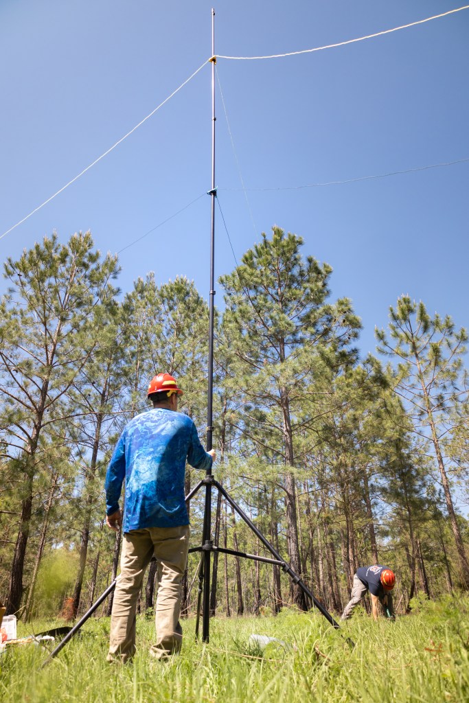 Two men in blue shirts with red hardhats mount equipment secure long, thin lines to support equipment on a tall tripod in a grassy field.