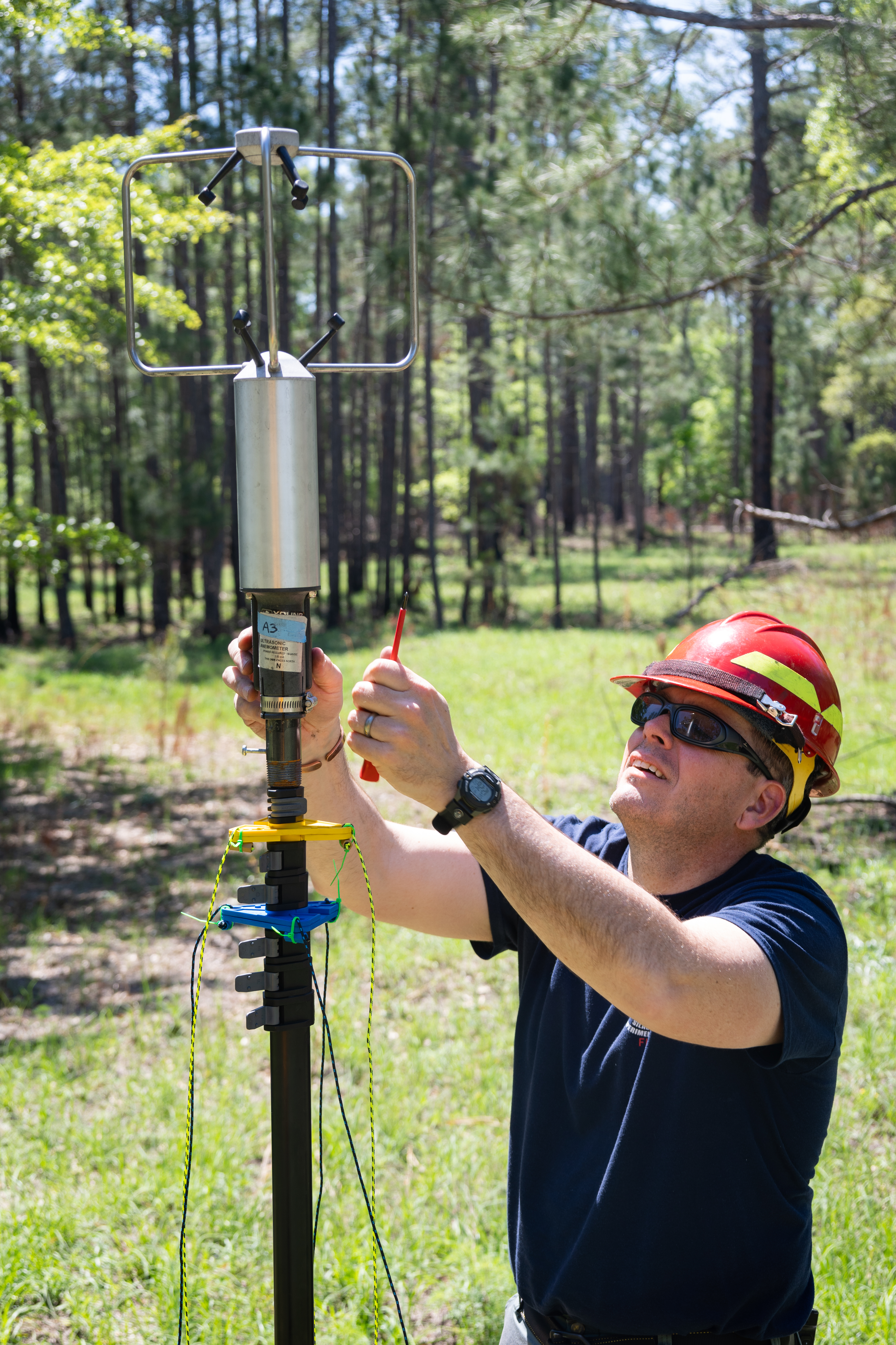 A man in a navy t-shirt wearing a red hard hat helmet and sunglasses uses a screwdriver to mount a silver cylindrical device onto a black pole in a grassy field.