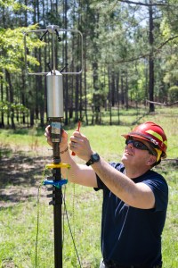 A man in a navy t-shirt wearing a red hard hat helmet and sunglasses uses a screwdriver to mount a silver cylindrical device onto a black pole in a grassy field.