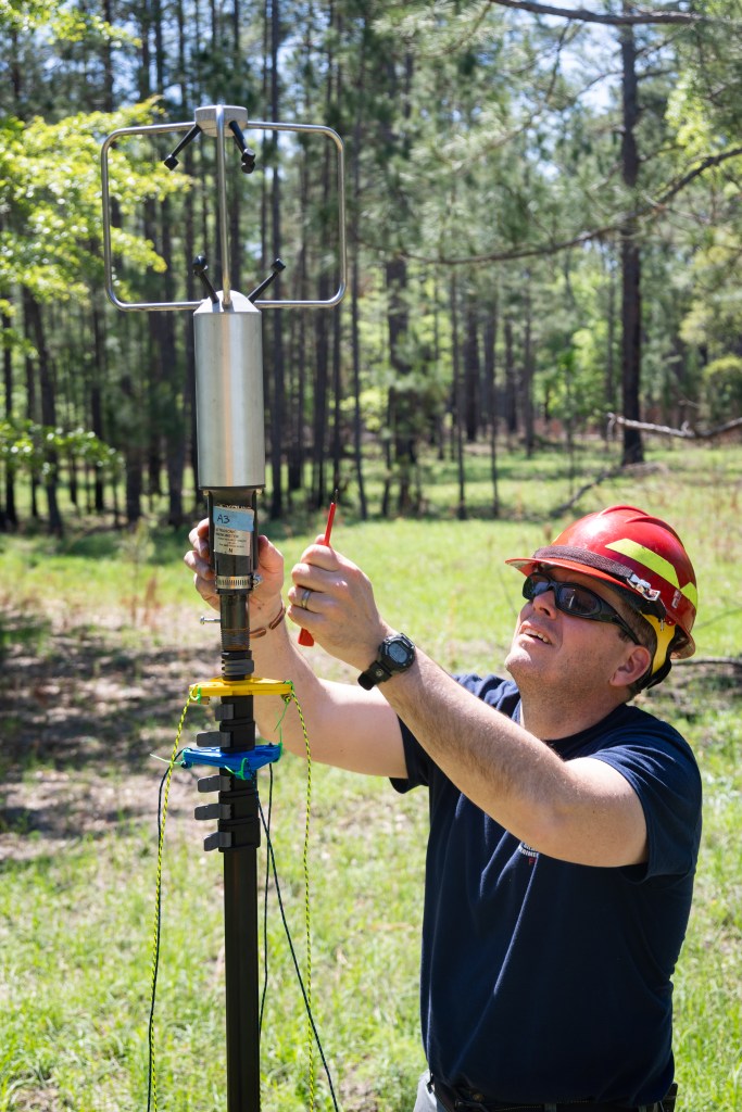 A man in a navy t-shirt wearing a red hard hat helmet and sunglasses uses a screwdriver to mount a silver cylindrical device onto a black pole in a grassy field.