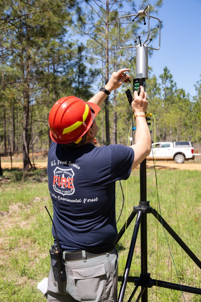 A man in a navy t-shirt wearing a red hard hat helmet and sunglasses uses a screwdriver to mount a silver cylindrical device onto a black pole in a grassy field.