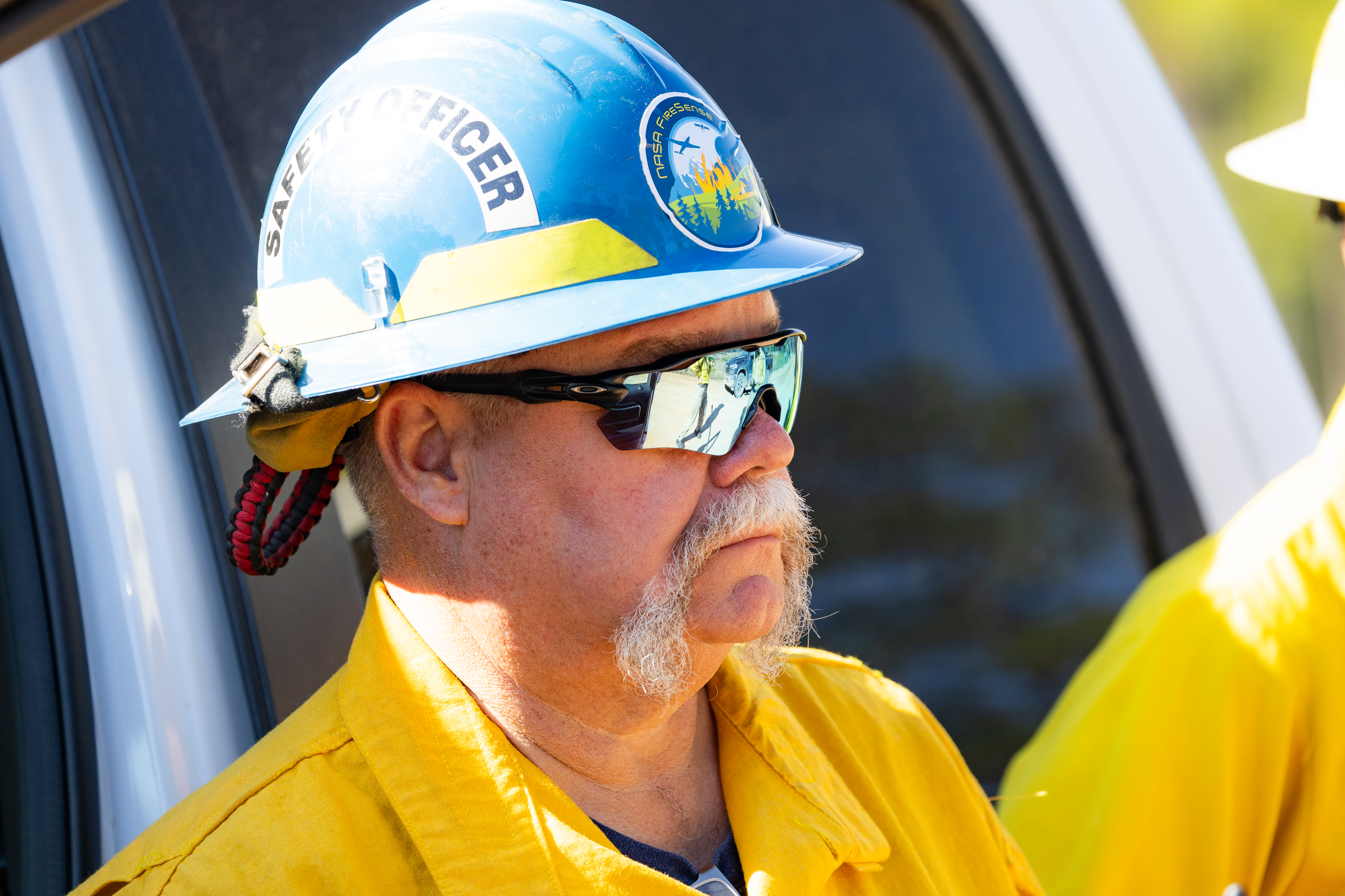 A man with a massive white mustache, wearing a bright yellow long-sleeved shirt, blue hard hat, and sunglasses looks to the right.