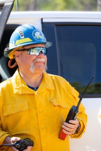 A man with a massive white mustache, wearing a bright yellow long-sleeved shirt, blue hard hat, and sunglasses hold two radios.