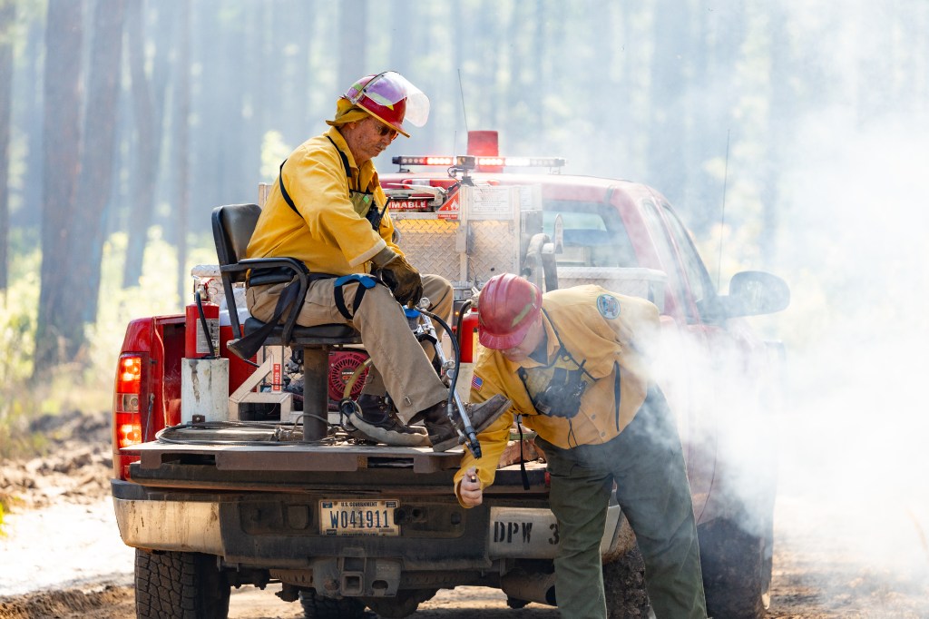 Two firefighters in a smoky environment; one sits in the bed of a red pickup truck holding a long thin device, the other holds the lighter to the base of the device.