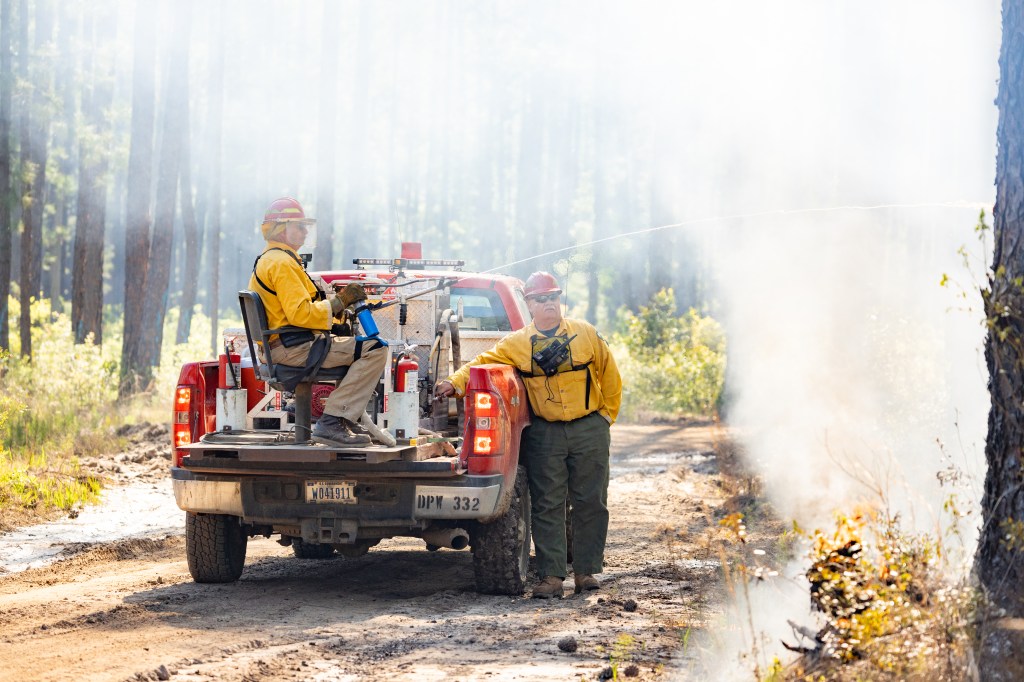 Two firefighters in a smoky environment; one sits in the bed of a red pickup truck holding a long thin device shooting an almost-clear substance into the trees, the other leaning against the side of the truck.