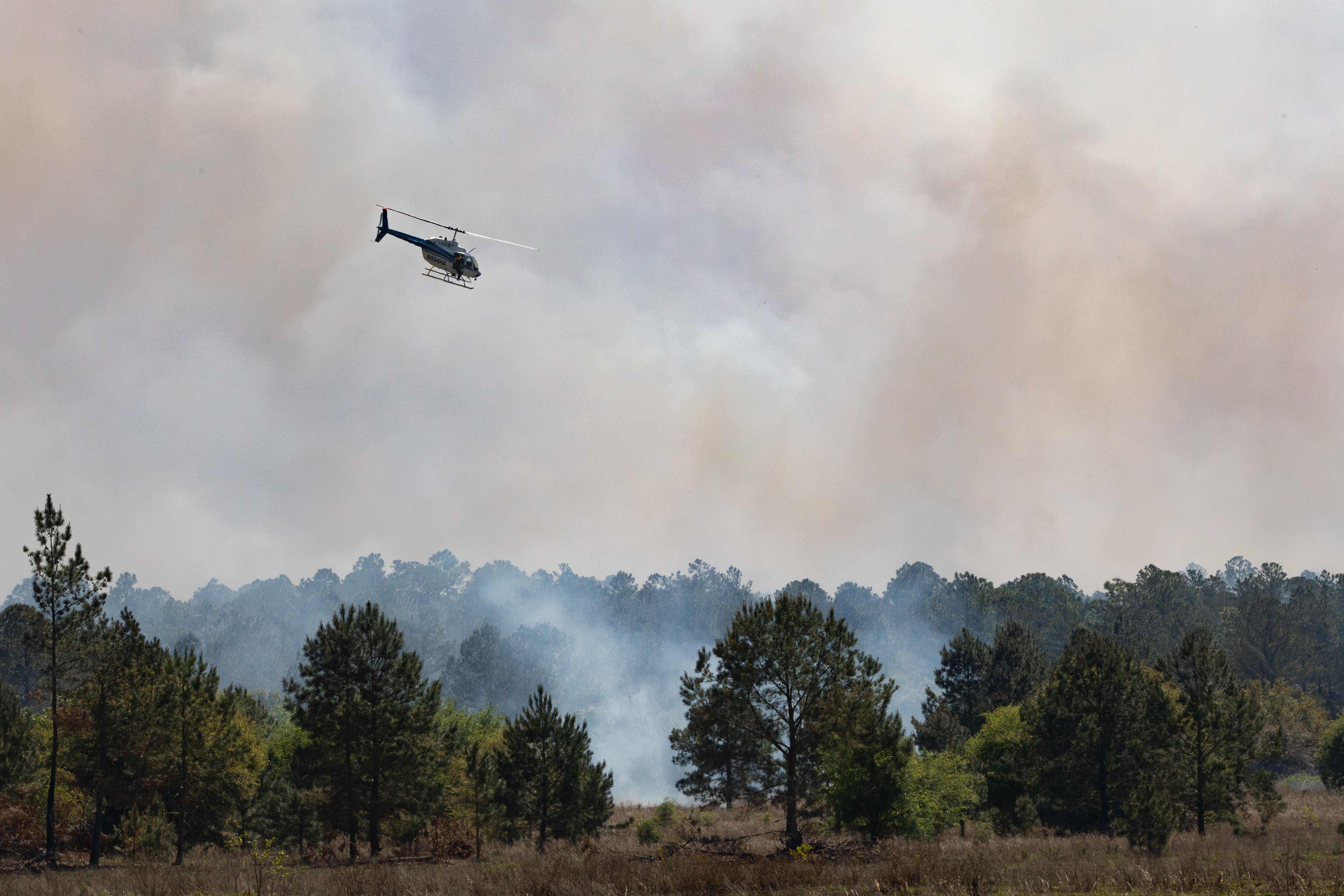 A helicopter looks small as it flies low over a forest, silhouetted against smoke.