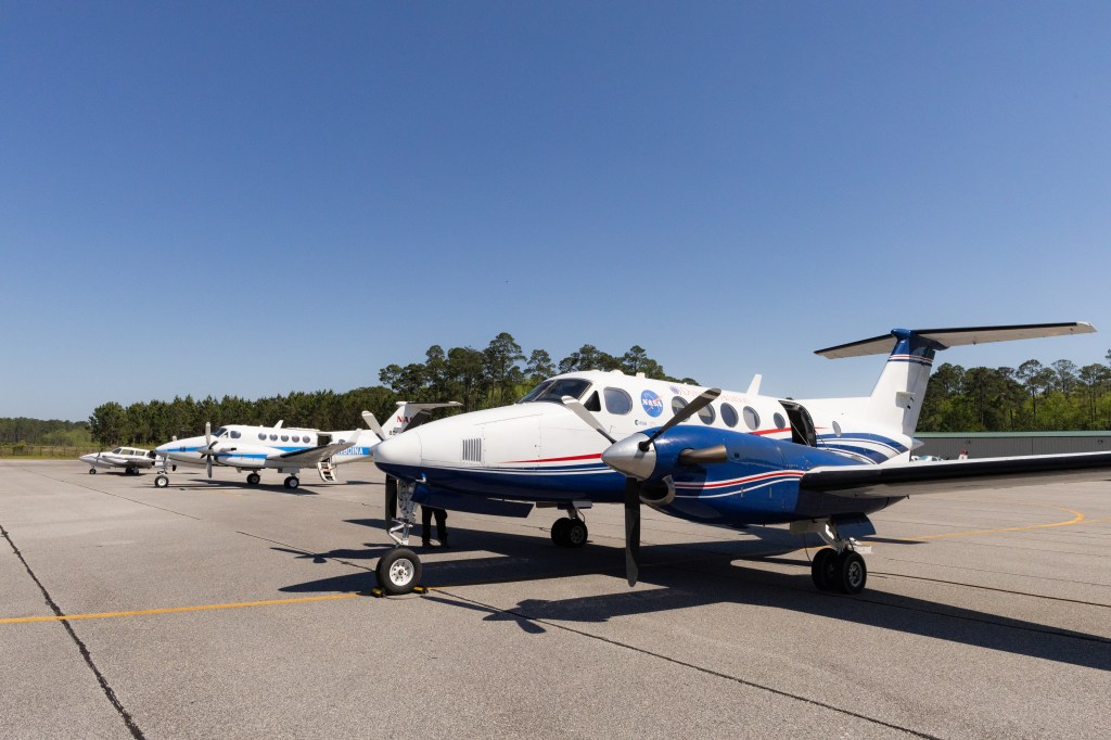 Three aircraft sit in a row stretching away from the camera on a gray tarmac, with blue sky in the background.