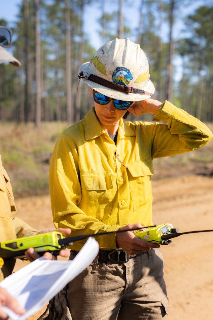 A woman wearing a yellow long-sleeved and white hard hat holds a yellow radio and looks at a paper held by someone off-camera.