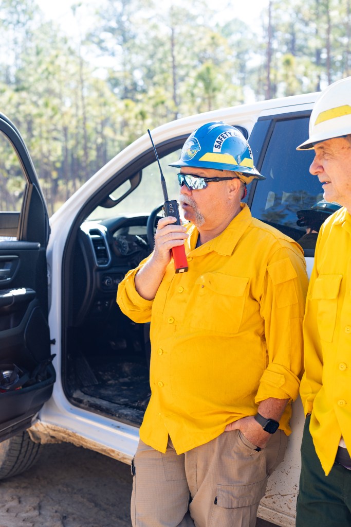 Two safety officers in yellow long-sleeved shirts and hardhats stand in front of white car in forest. The one on the left speaks into a red radio.