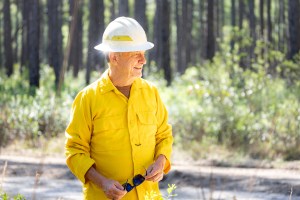A man in a yellow long-sleeved and white hard hat holds sunglasses in his hands as he smiles, while looking to his left.