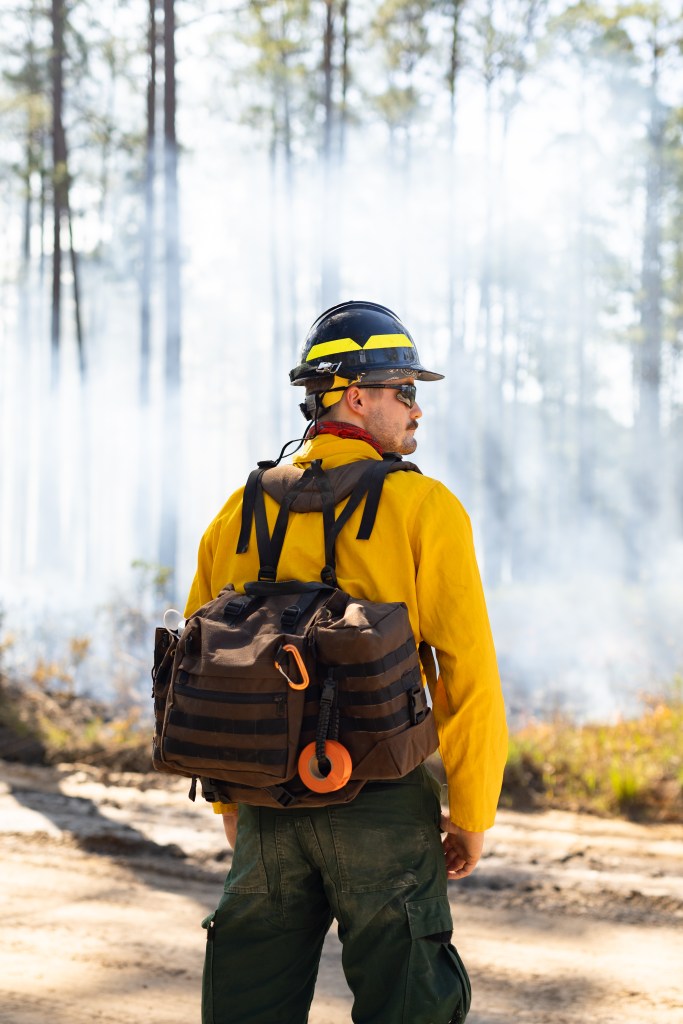 A firefighter in a yellow long-sleeved shirt, khaki green pants, black hard hat and brown backpacks stands with his back to the camera, looking at a smoke-filled forest.