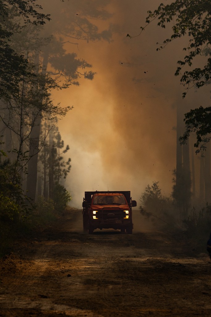 A red pickup truck with headlights on in barely visible, driving down a dirt road amidst a dark smokey forest.