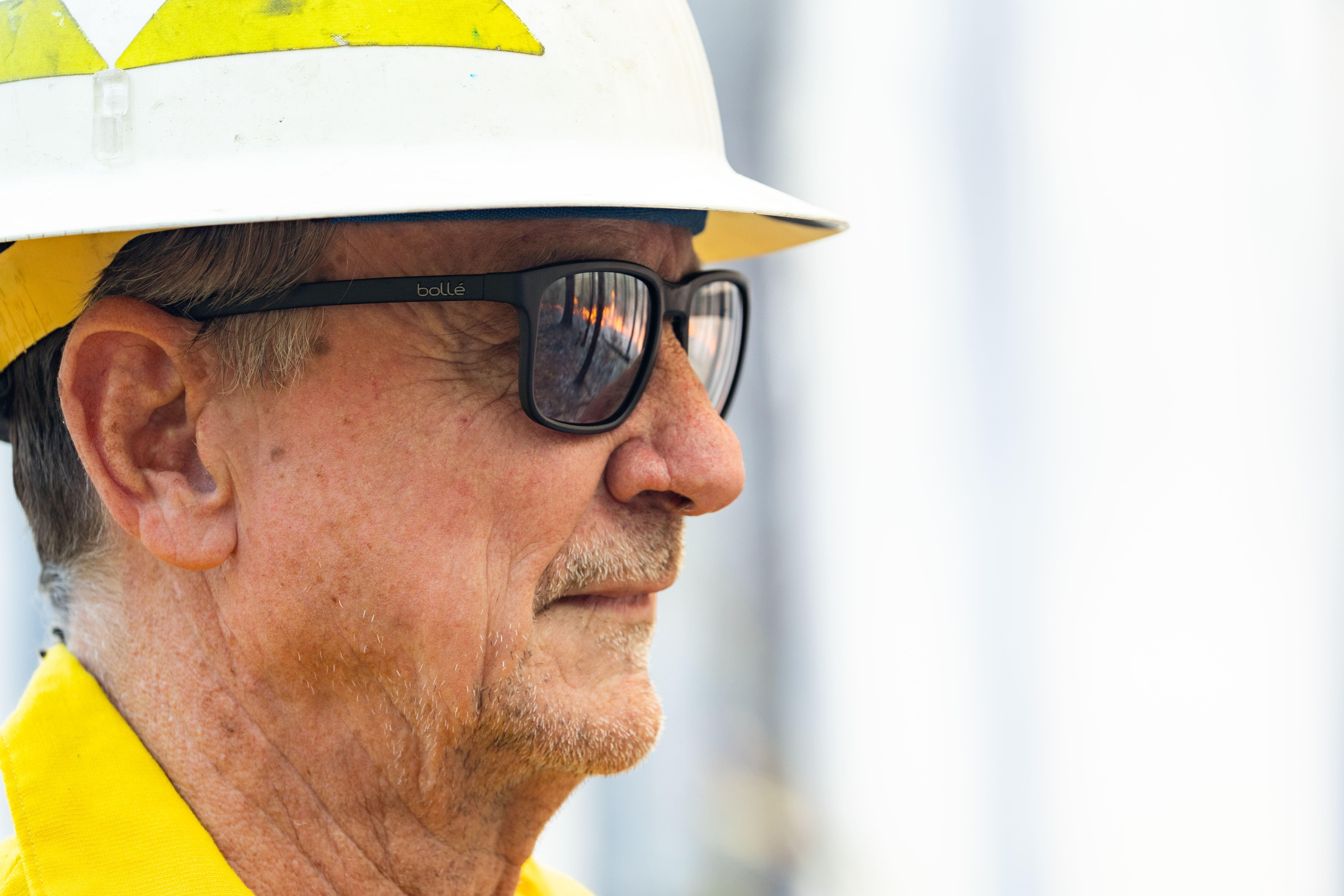 A closeup shot of a man with stubble, wearing a white hard hat and black sunglasses. Fire and the outline of trees can be clearly seen reflecting in the sunglasses.