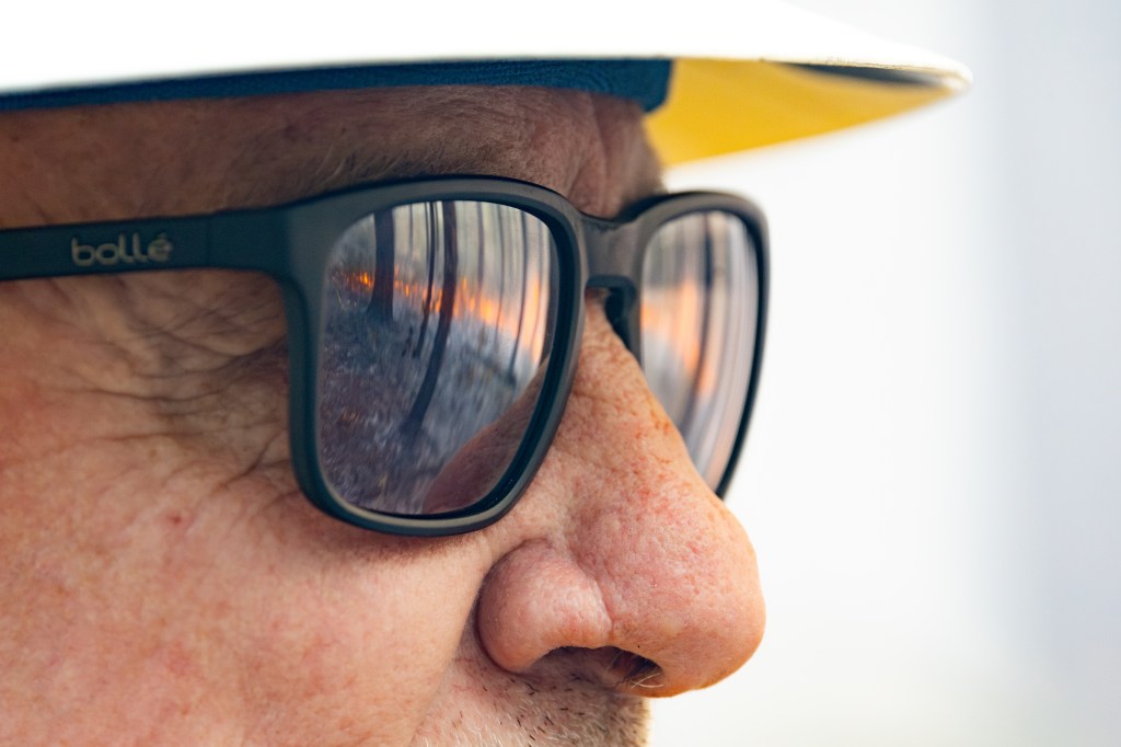 A closeup shot of a man with stubble, wearing a white hard hat and black sunglasses. Fire and the outline of trees can be clearly seen reflecting in the sunglasses.