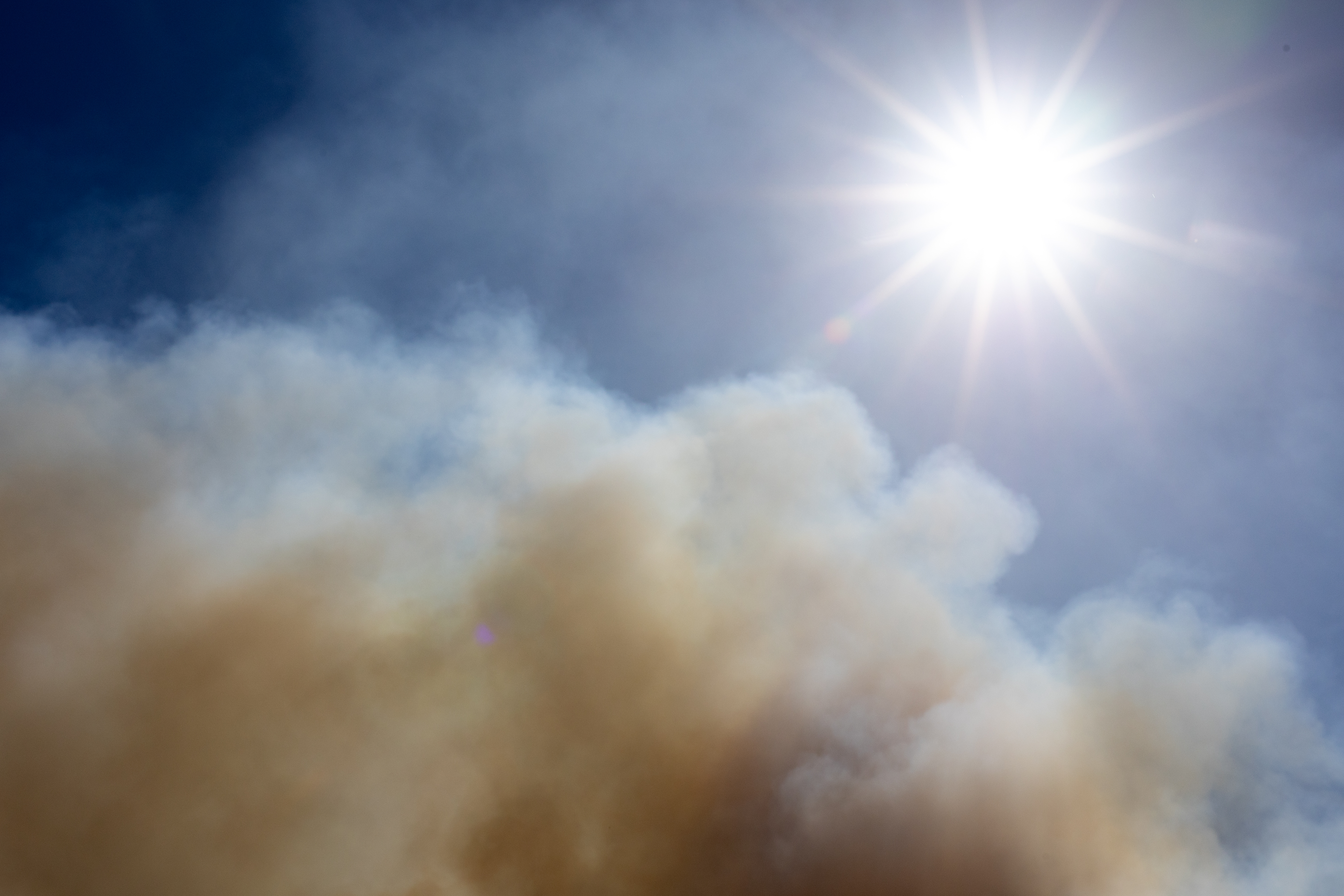 Bright sun shines in a blue sky, as a dark smokey cloud rises into the frame from the bottom left.