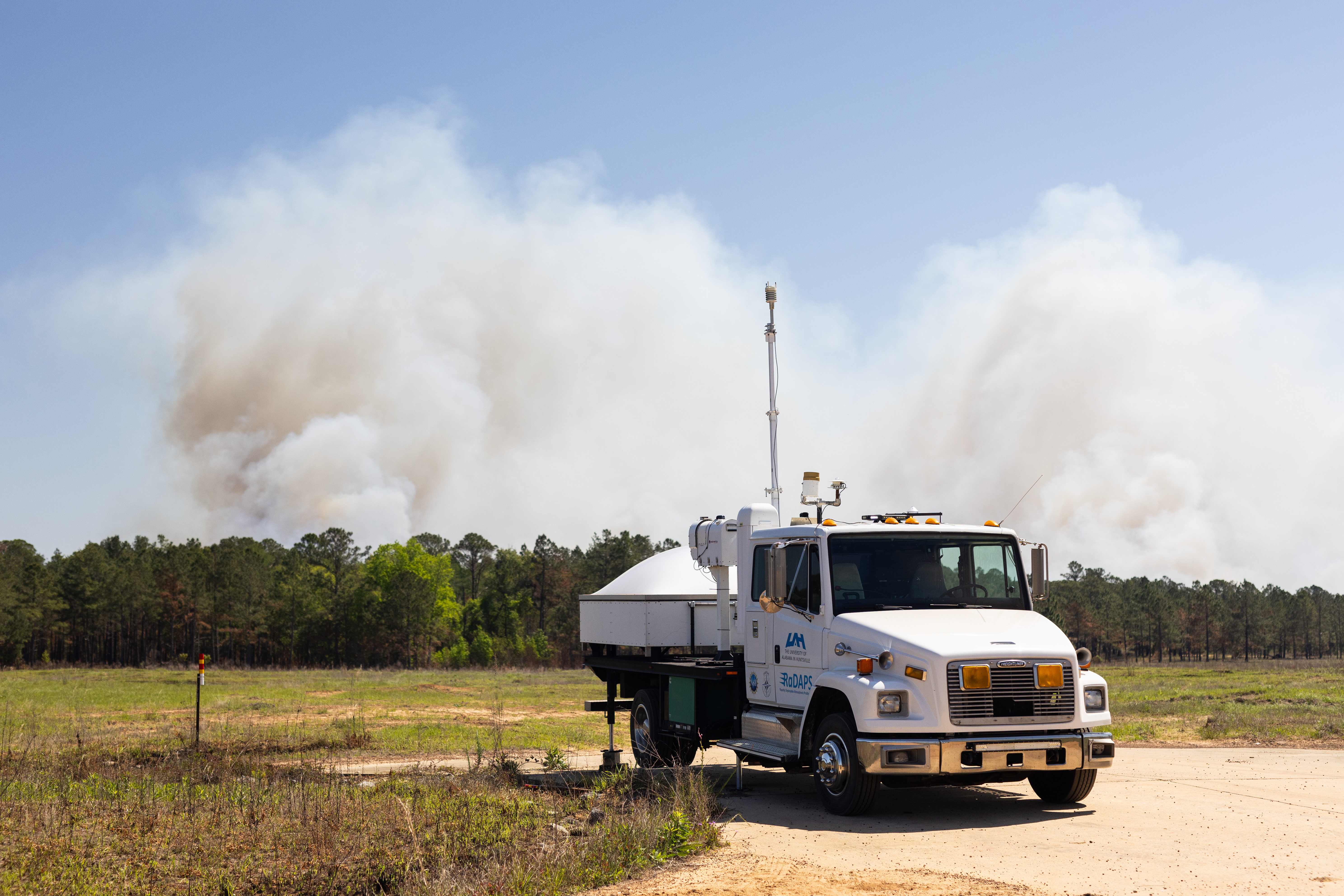 A white truck sits facing the camera in a clearing as a massive plume of white smoke rises into blue sky in the distance.