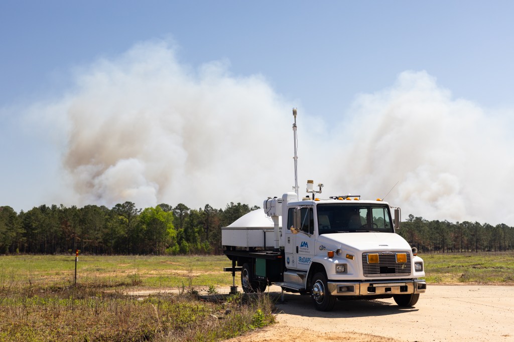 A white truck sits facing the camera in a clearing as a massive plume of white smoke rises into blue sky in the distance.