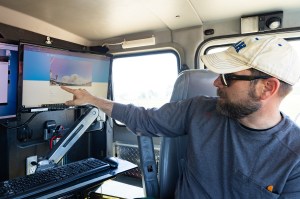 A man in a white hat and blue shirt points to smoke plume on monitor from inside a vehicle.