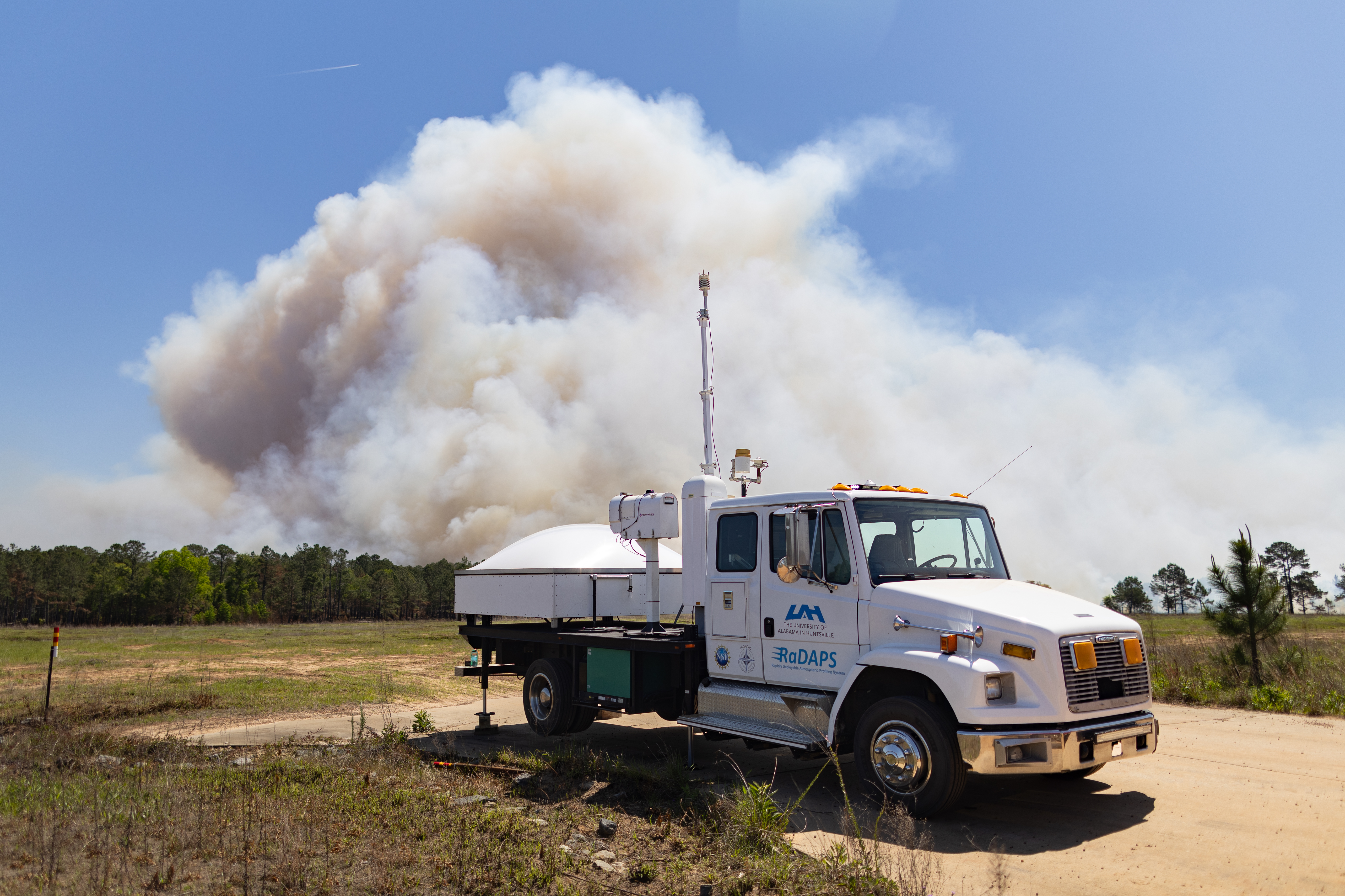 A white truck with a radar on the back sits in a clearing as a massive plume of white smoke rises into blue sky in the distance.