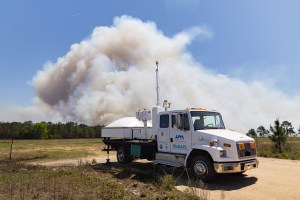 A white truck with a radar on the back sits in a clearing as a massive plume of white smoke rises into blue sky in the distance.