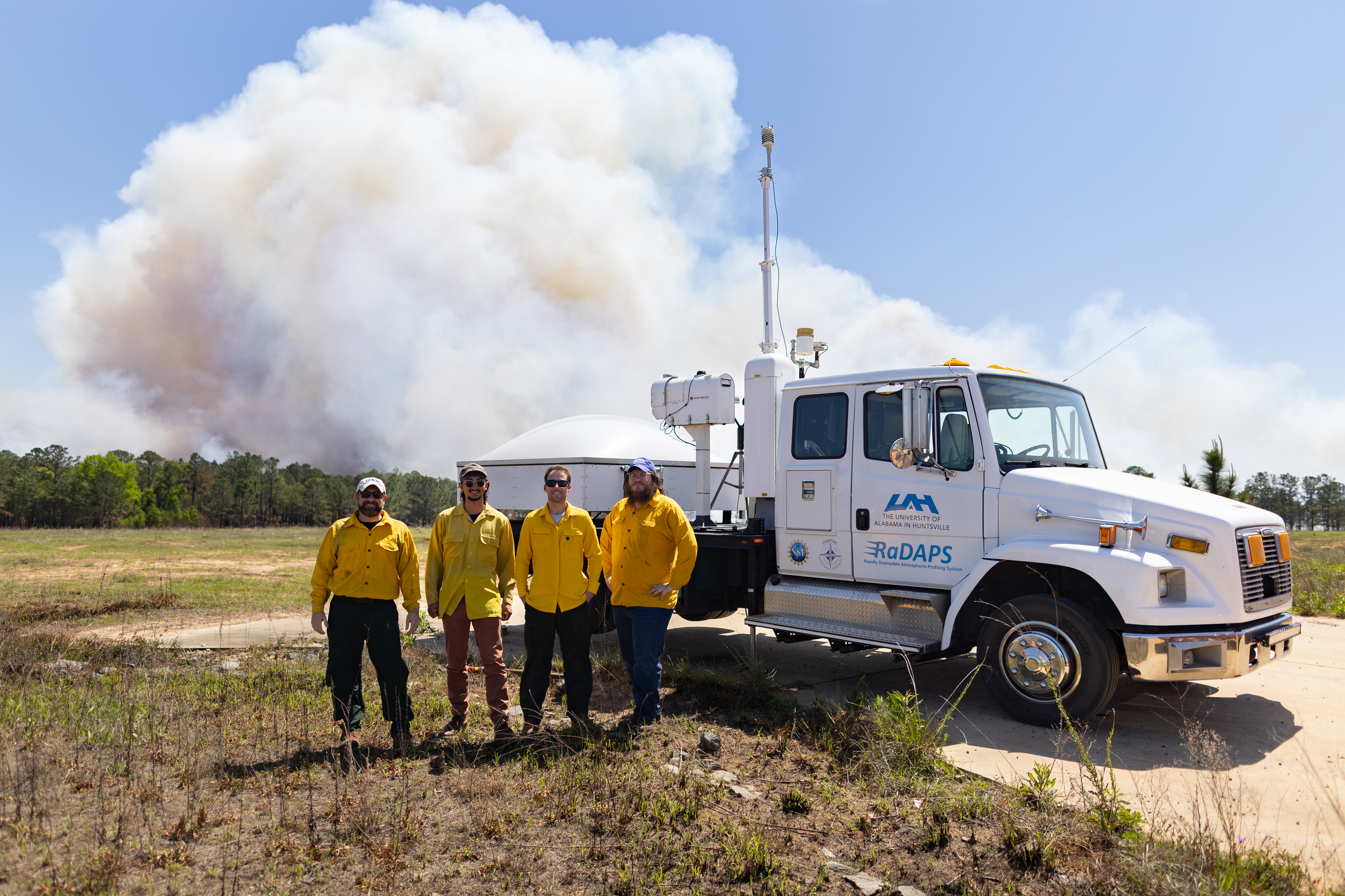 Four men in yellow long-sleeved shirts pose in front of a white truck with a radar on the bed, while a white smoke plume drift into blue sky behind them.