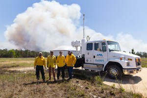 Four men in yellow long-sleeved shirts pose in front of a white truck with a radar on the bed, while a white smoke plume drift into blue sky behind them.