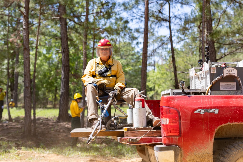 A firefighter with a red helmet sits in a seat on the bed of a red pickup truck as it drives through the forest, holding a long cylindrical device.