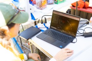 A scientist looks at a data visualization, white spots on a black background, on a laptop sitting on a on white table that also holds a notebook and computer cords.