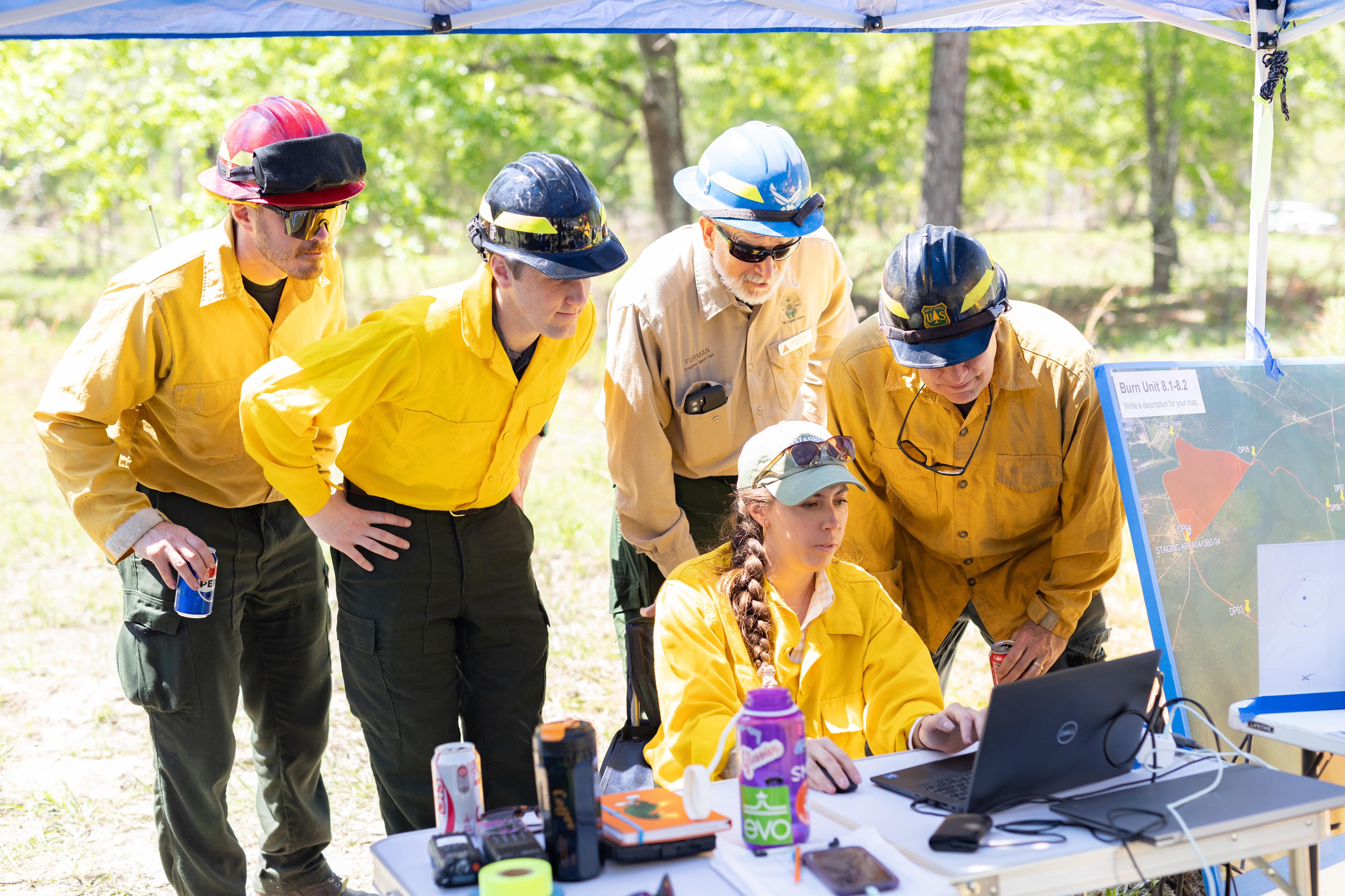 Four men in yellow long-sleeved shirts and hardhats gather around a woman with a brown braid over her shoulder, sitting at a table with a laptop. Everyone is looking at the laptop.