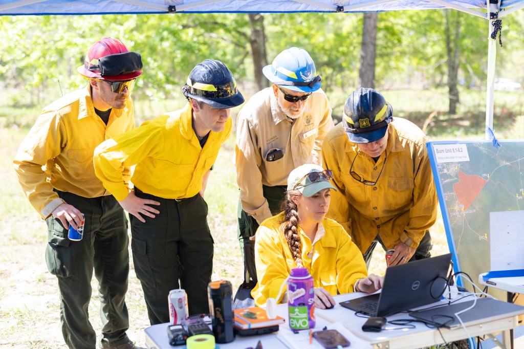 Four men in yellow long-sleeved shirts and hardhats gather around a woman with a brown braid over her shoulder, sitting at a table with a laptop. Everyone is looking at the laptop.