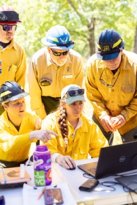 Four men in yellow long-sleeved shirts and hardhats gather around a woman with a brown braid over her shoulder, sitting at a table with a laptop. Everyone is looking at the laptop.