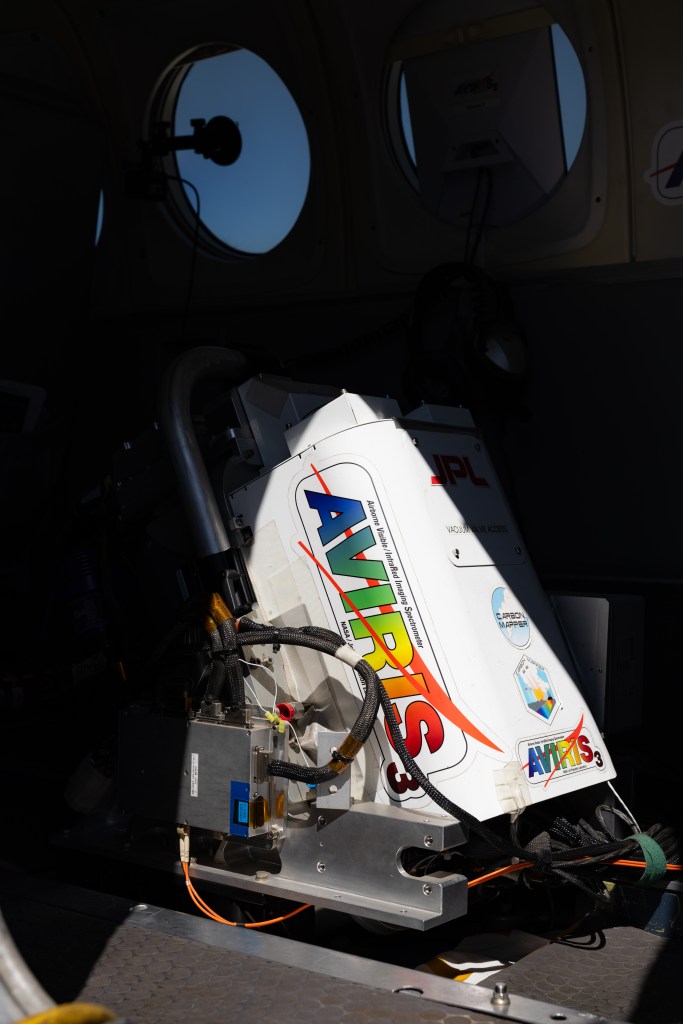An instrument that looks like a white box mounted at an angle has the word AVIRIS on the side in rainbow font, partially illuminated by the sun.