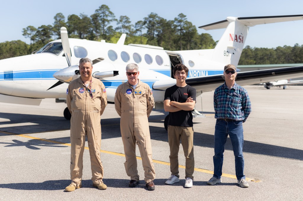 Two pilots in tan flight suits and two instrument specialists stand in front of a white NASA Armstrong B-200 aircraft on gray tarmac.